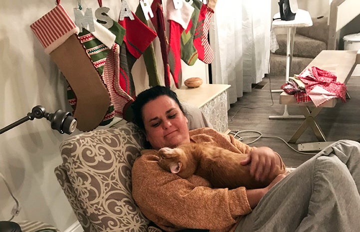 Sara sitting on a chair holding Dan the orange cat in her lap with Christmas stocking hanging up behind them