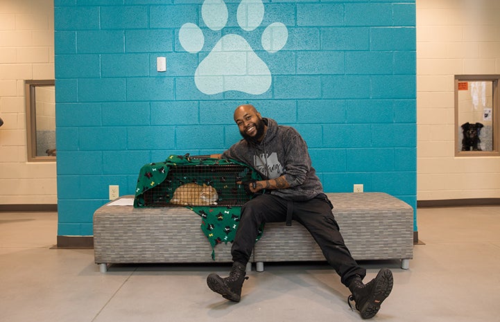 Sterling “Trap King” Davis with a cat in a humane trap, sitting on a bench in front of a blue wall with a big paw print