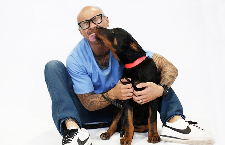 Man with tattoos hugging a dog who is giving him a kiss on the face