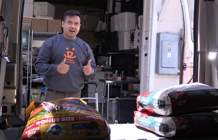 Volunteer Christian Ordonez wearing a Best Friends sweatshirt giving a thumbs up from behind some large bags of dog food