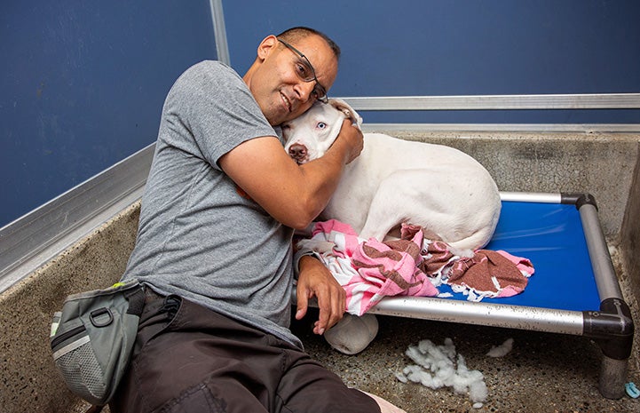 Volunteer Robert Lobo lying on a dog bed hugging Delilah the dog in a kennel