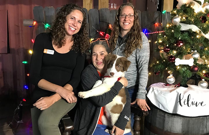 Deya Galvan holding a puppy with Amy Kohlbecker and Whitney Bliton, next to a small Christmas tree