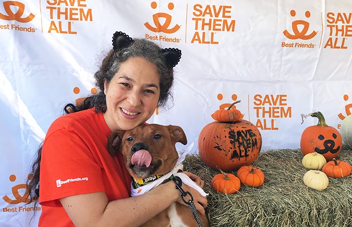 Volunteer Deyra Galvan wearing cat ears and hugging a dog who is licking his lips, next to some pumpkins