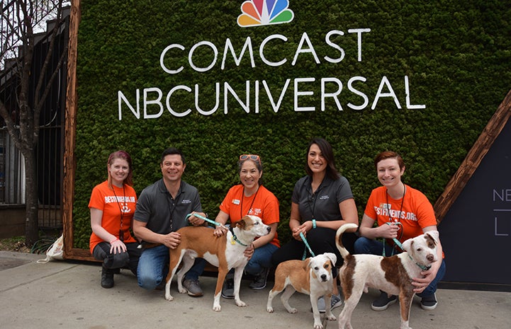 Volunteer Deya Galvan with other people and three dogs in front of a Comcast NBCUniversal sign