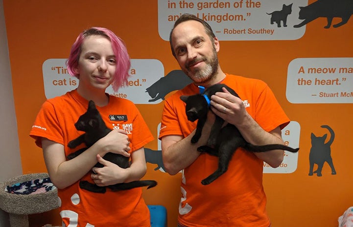 Volunteers Bella and Justin Brodie each holding black kittens while standing in front of an orange wall with graphics