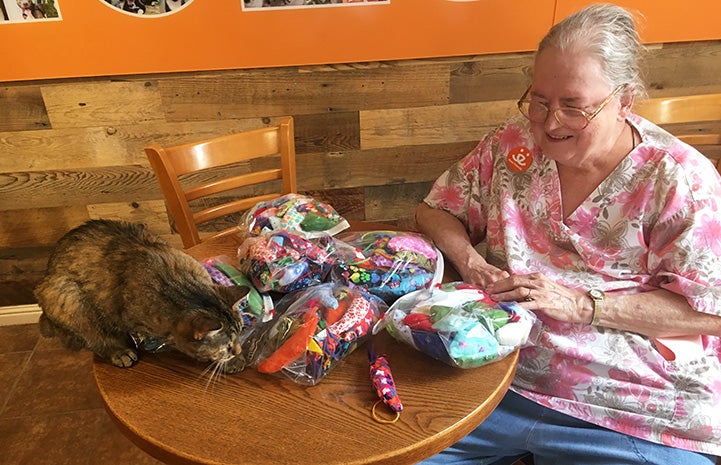Marilyn Mott sitting at a table holding the bags of mouse-shaped cat toys she made and donated, while torbie cat is ready to pounce on one