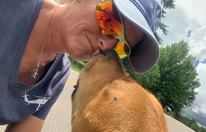 Becky McCrea, wearing sunglasses and a hat, getting a kiss from a big brown dog