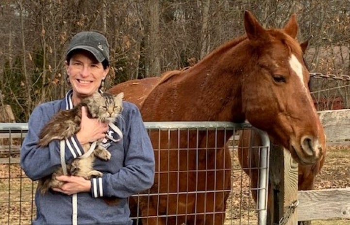 Kimberly holding Squash the cat in front of Oliver the horse