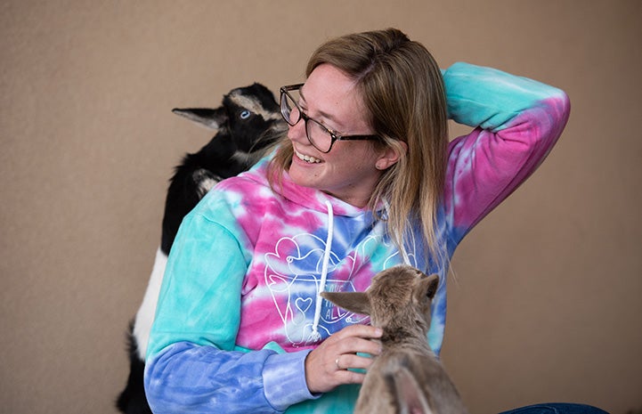 Smiling person wearing a tie-dye shirt reaching up to the baby goat at her shoulder and petting one in front of her