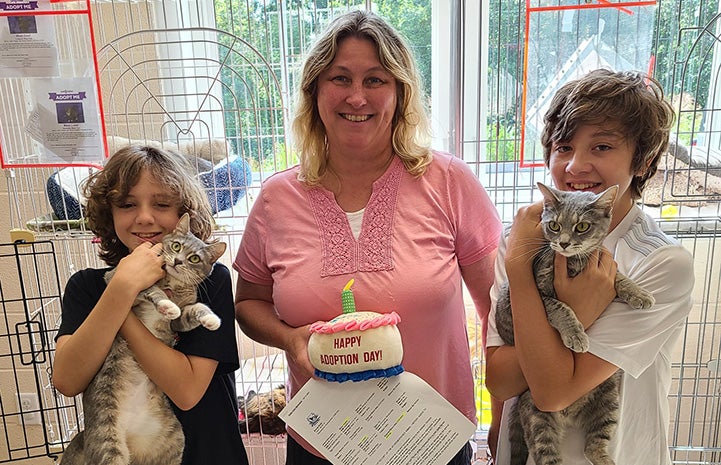 Woman holding a stuffed "Happy Adoption Day" cake and two kids, who are each holding a cat