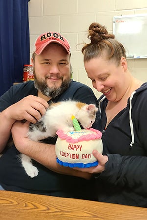 Smiling couple holding a cat and a stuffed "Happy Adoption Day" cake