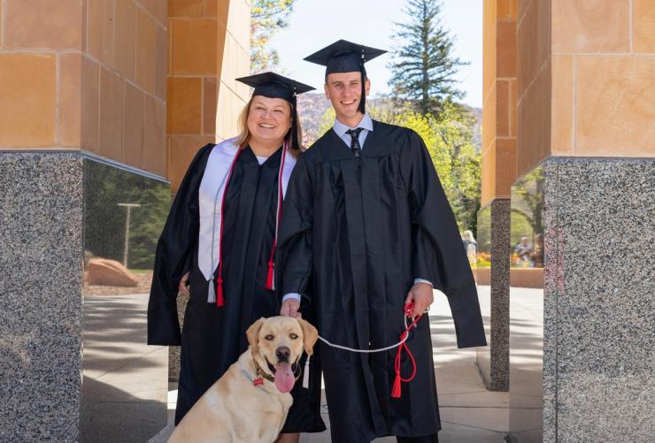 Two graduates from SUU wearing caps and gowns with a dog on a leash
