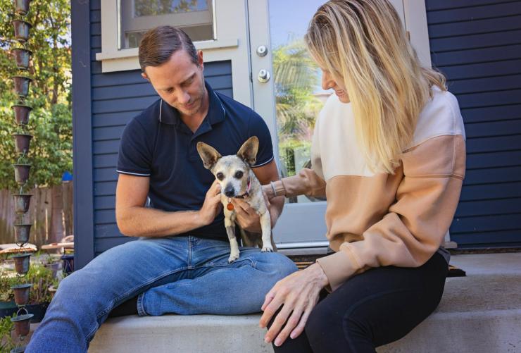 Two people sitting outside with a small dog on stairs leading into a hous