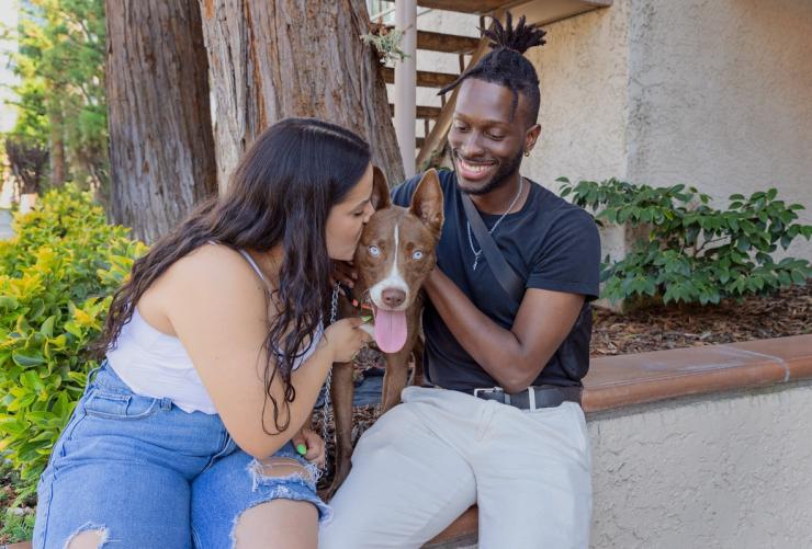 Two smiling people sitting outside with a dog between them