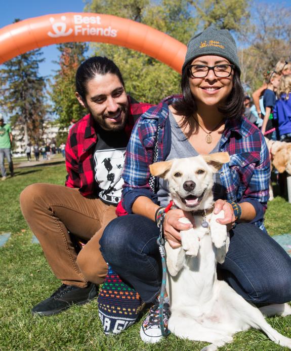 Two happy people sitting with a dog in the grass at an outdoor event