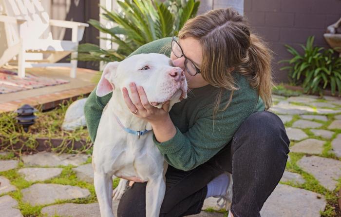 Person kneeling down next to a dog and kissing the side of their face