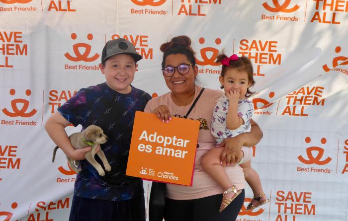 Family adopting a puppy on front of a Best Friends backdrop holding a sign that says, Adoptar es amar