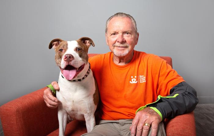 Smiling person in orange Best Friends volunteer T-shirt sitting in a chair next to a smiling brown and white dog