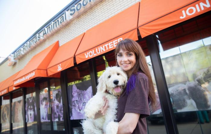 Smiling person holding a small white dog in front of a Best Friends regional location