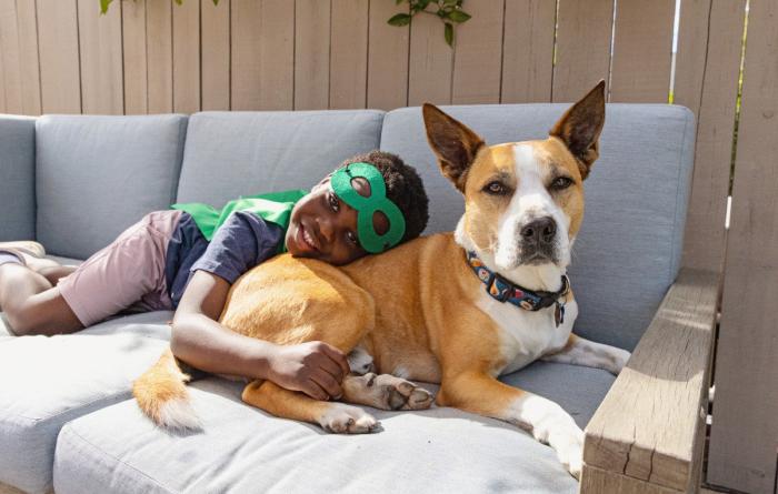 Child wearing a green superhero mask and cap lying on the back of a dog on a couch