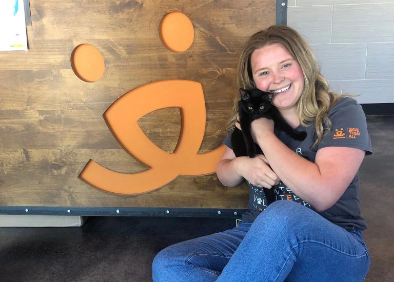Woman holding a black cat sitting on the ground next to a Best Friends logo on the wall
