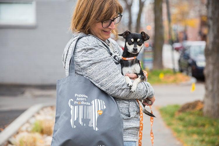 Person smiling and holding a small dog, along with a Puppies Aren't Products Best Friends bag