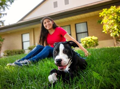 Smiling person sitting on the grass in front of a house with a happy dog