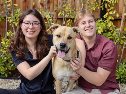 man and woman sitting outside by wooden fence with large light brown dog