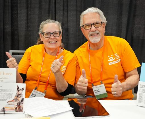 Two volunteers in orange volunteer shirts giving thumbs up at conference booth table