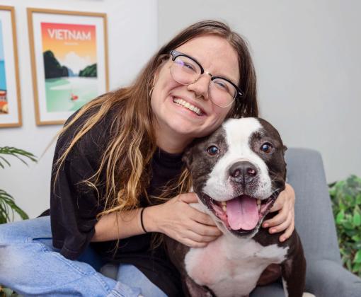 Smiling person hugs a pit bull mix on a gray chair indoors, both facing the camera