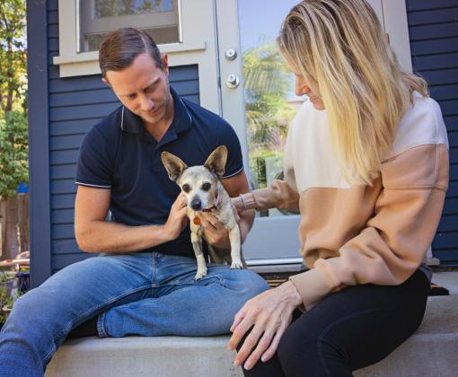 Two people sitting outside with a small dog on stairs leading into a hous