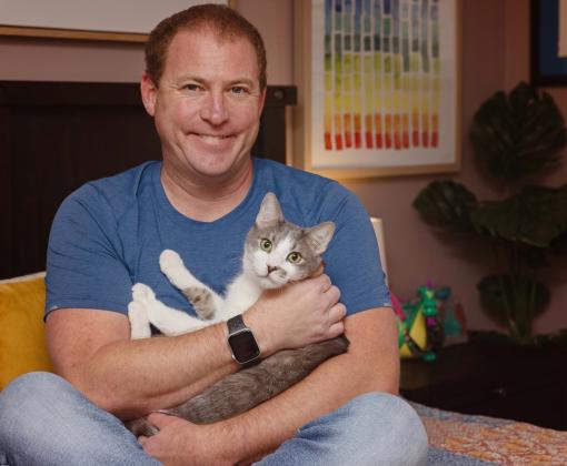 Smiling person cradling a cat in his arms while sitting on a bed in a home