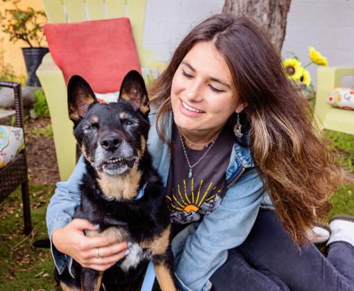 Smiling person sitting on the ground next to a dog while hugging it