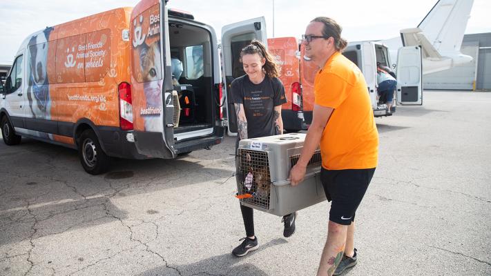 People carrying a dog in a crate from a Best Friends transport van