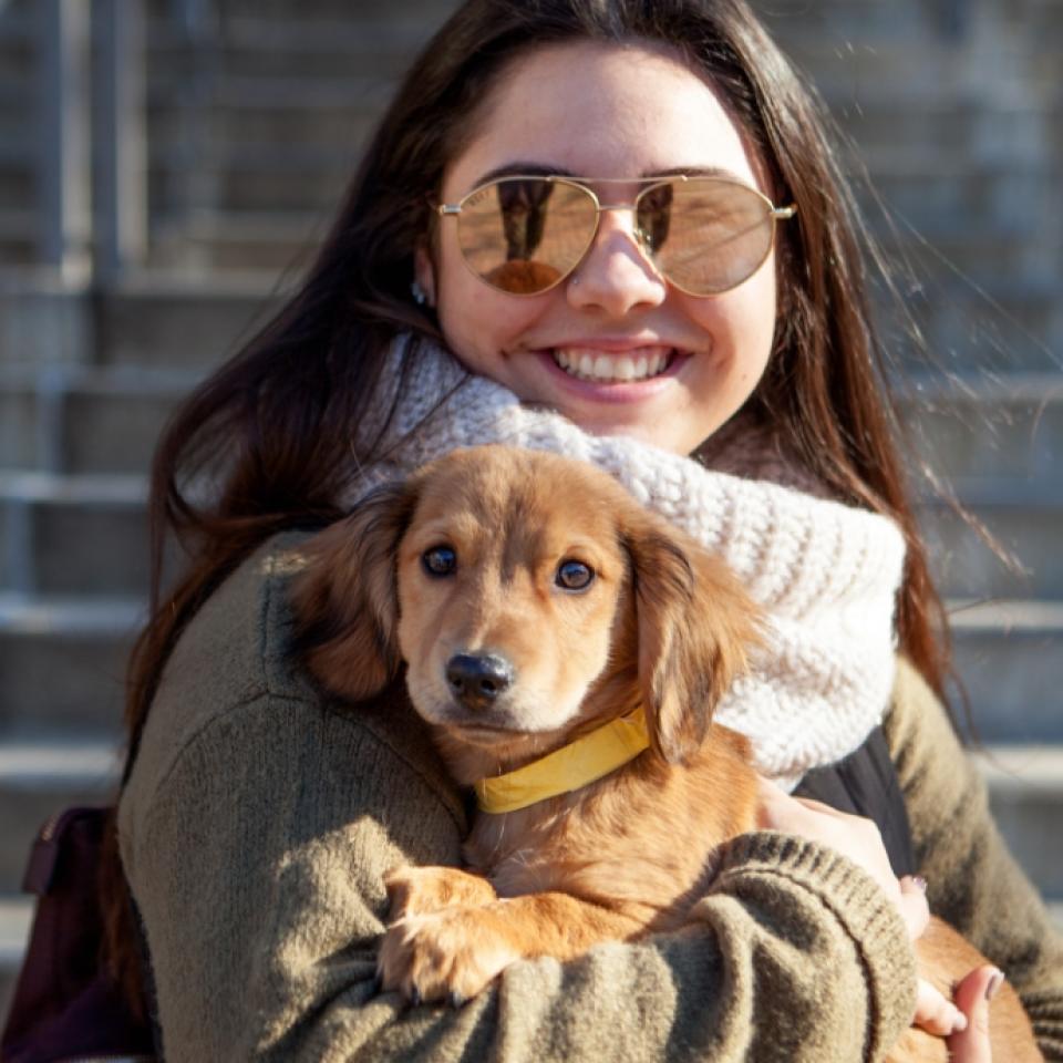 Smiling person wearing sunglasses and a sweater holding a small brown puppy