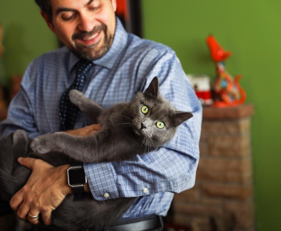 Smiling man cradling a gray cat in his arms like a baby, in front of a fireplace mantel