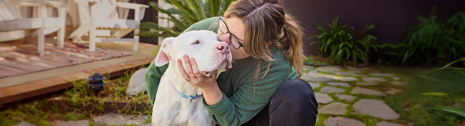 Person kneeling down next to a dog and kissing the side of their face