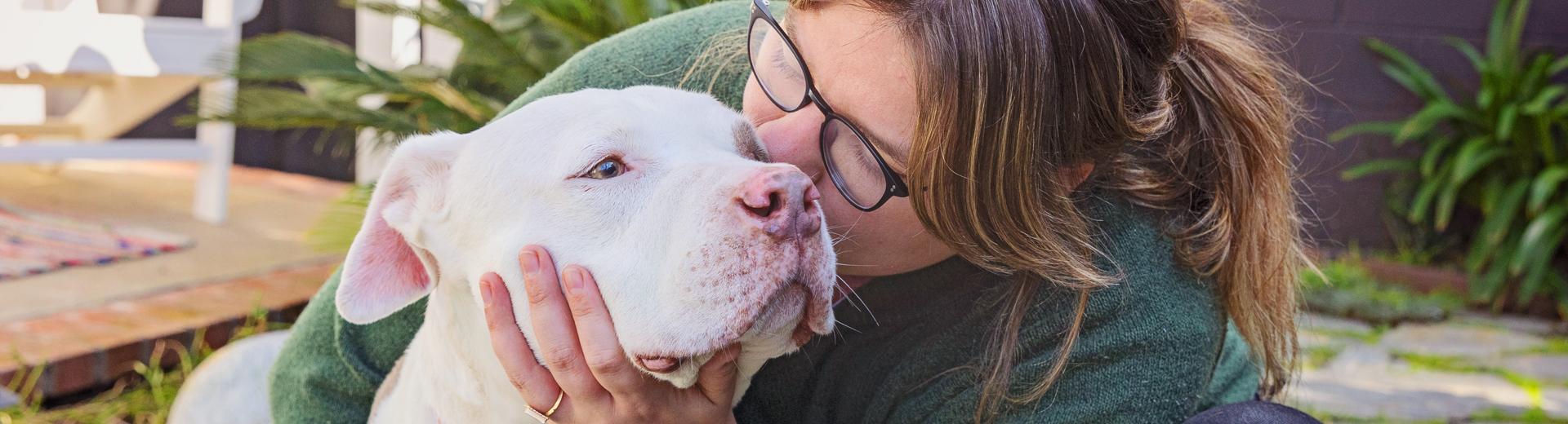 Person kneeling down next to a dog and kissing the side of their face