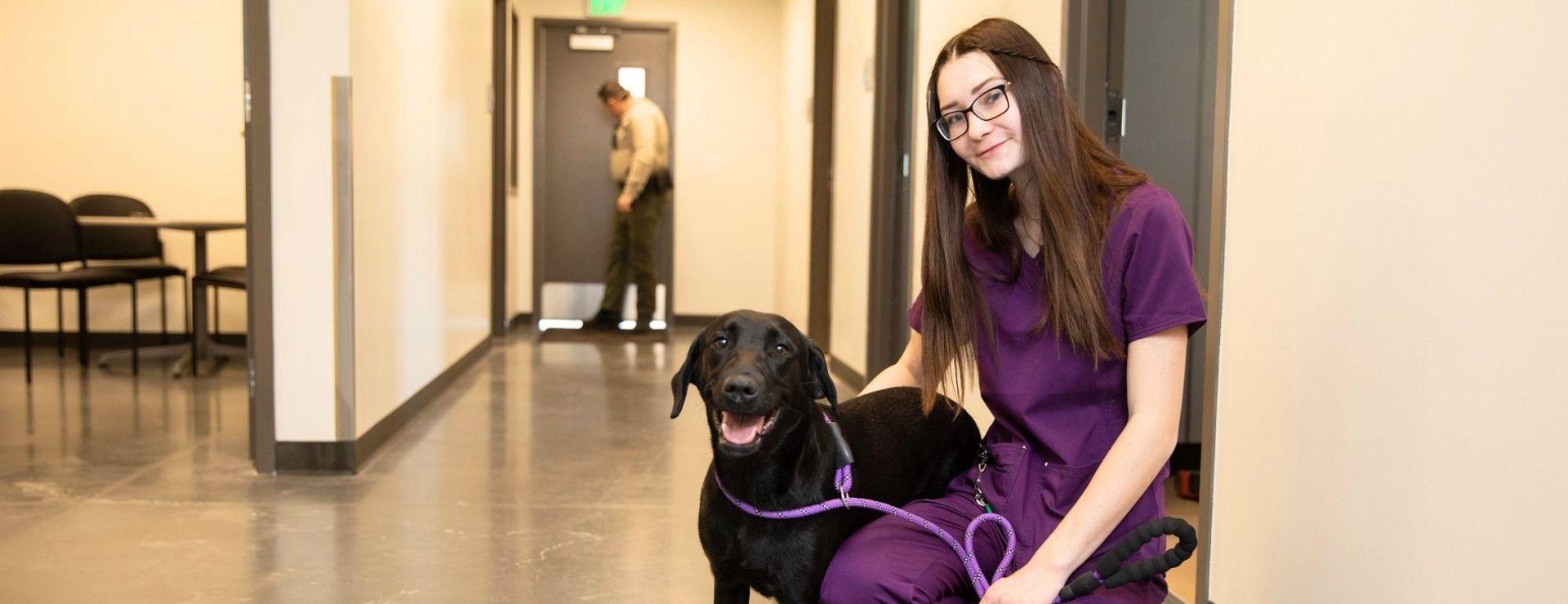 Woman with long brown hair in purple scrubs kneeling down with a black dog
