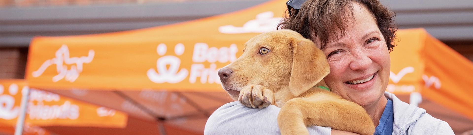 Smiling woman holding brown dog close in front of adoption event tent