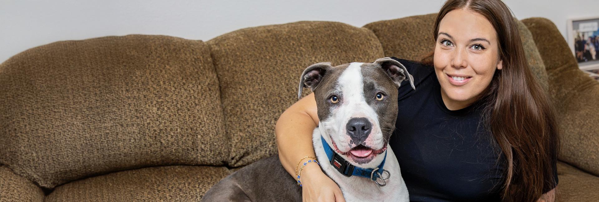 Smiling person with her arm around a happy gray and white pit bull terrier, both on a couch