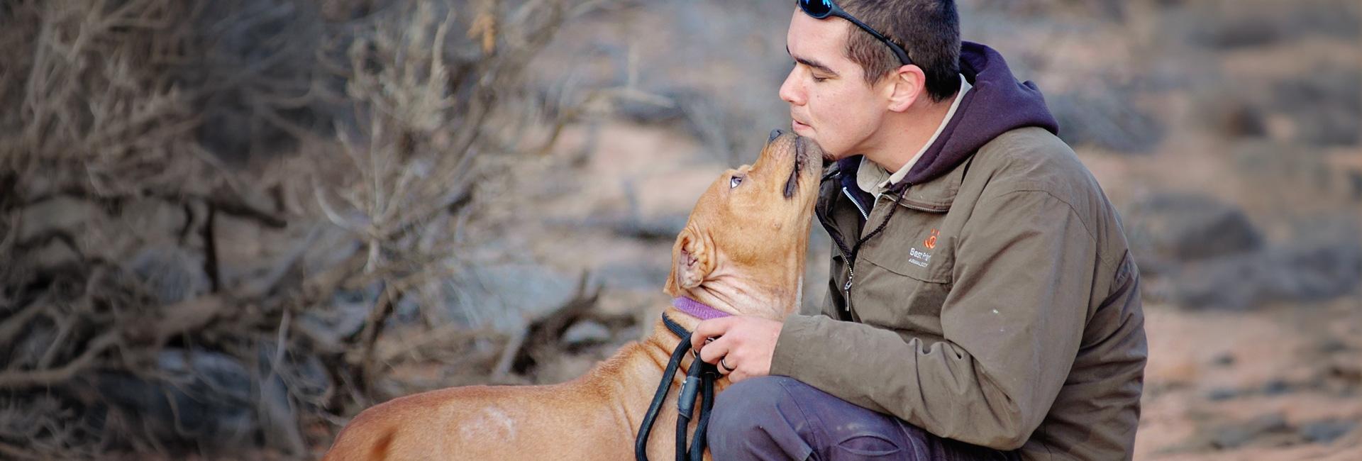 Man getting kiss from brown pit bull terrier in canyon setting