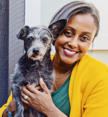 Smiling person wearing bright yellow and holding a small, senior dog next to a gray wall