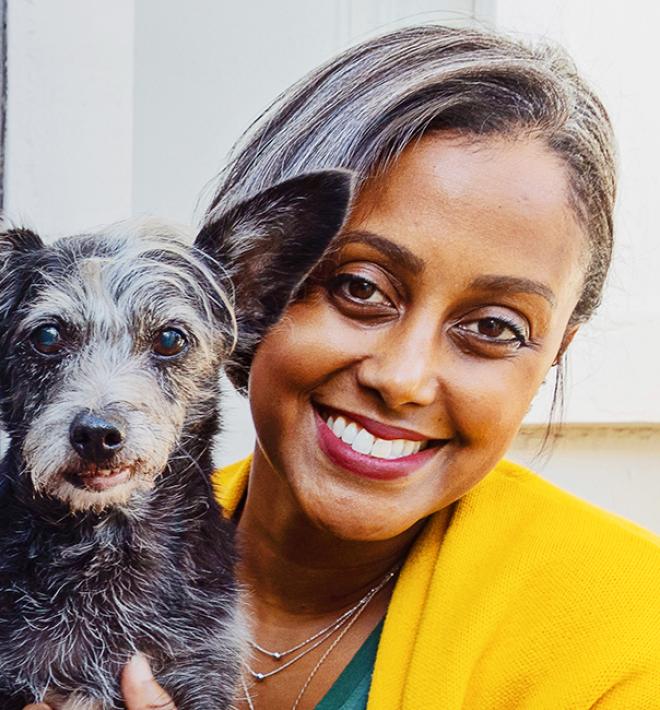 Smiling person wearing bright yellow and holding a small, senior dog next to a gray wall