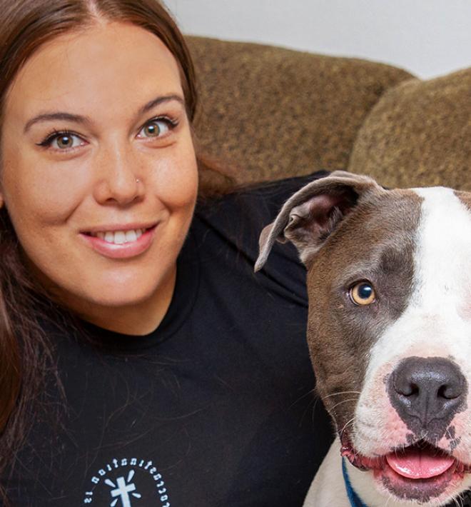 Smiling person with her arm around a happy gray and white pit bull terrier, both on a couch