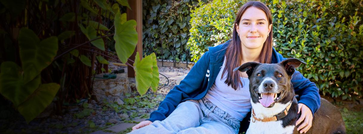 Smiling person sitting next to a dog in front of a green hedge