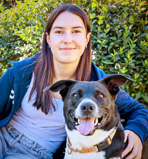 Smiling person sitting next to a dog in front of a green hedge