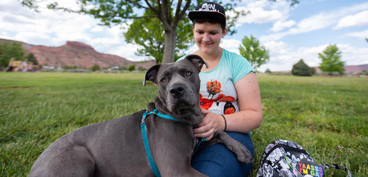 Smiling person with a big gray dog in the person's lap, sitting outside in the grass