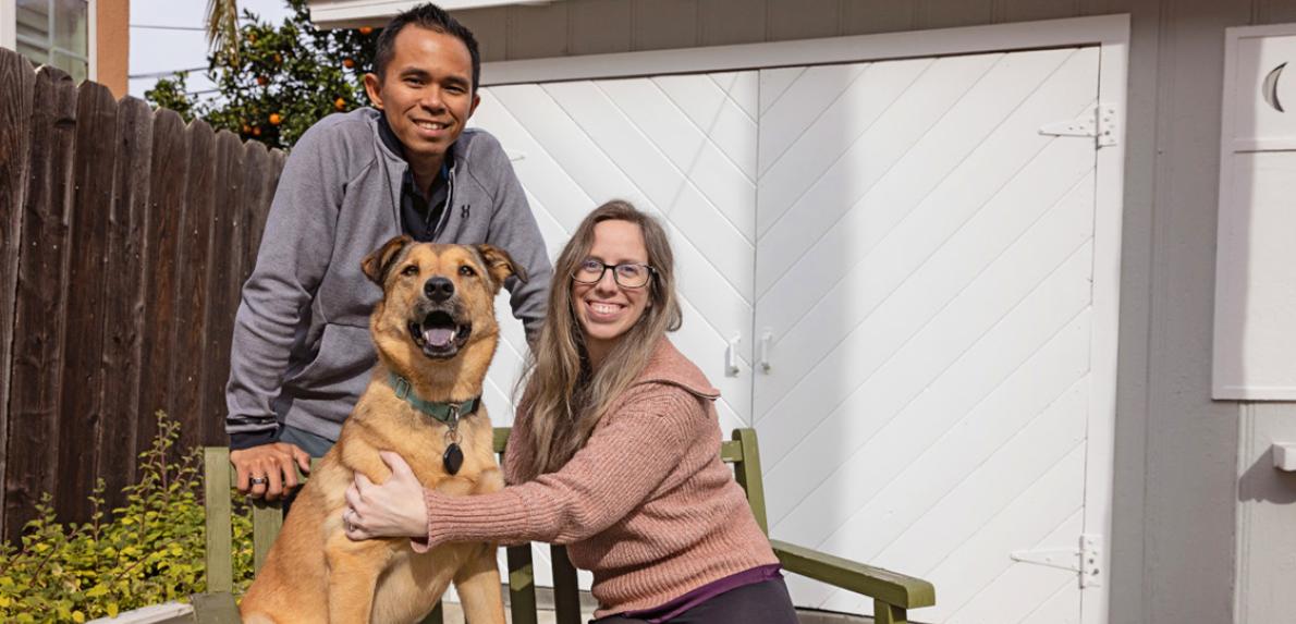 Two people with a dog between them, on and behind a bench, in front of double doors of a shed