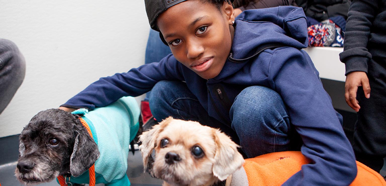 Young person crouched down with each arm around a small senior dog earing a coat